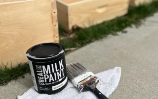 Can of plastic-free paint with brush on cloth beside unfinished wooden boxes outdoors.