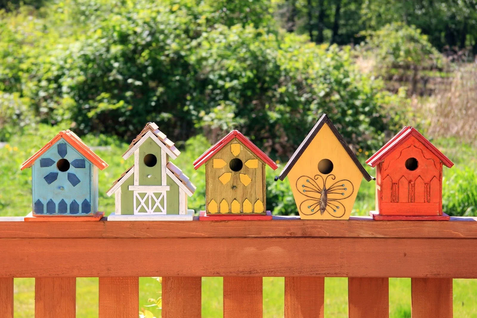 A row of five colorful wooden birdhouses sitting on a wooden deck railing with green foliage in the background.
