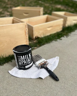 Can of plastic-free paint with brush on cloth beside unfinished wooden boxes outdoors.