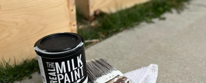 Can of plastic-free paint with brush on cloth beside unfinished wooden boxes outdoors.
