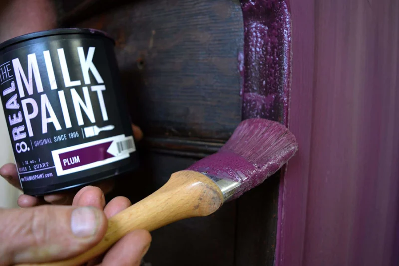 Man holding brush and applying eco-friendly paint in a deep plum color to wooden furniture beside an open paint can.